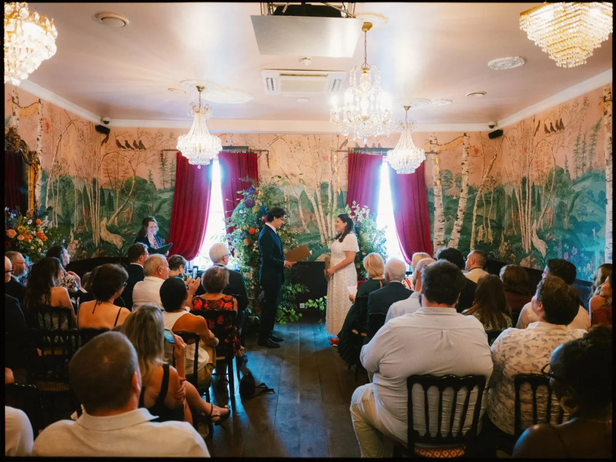Maya and Lukas face each other holding papers during the ceremony, guests seated beneath chandeliers at The Bell