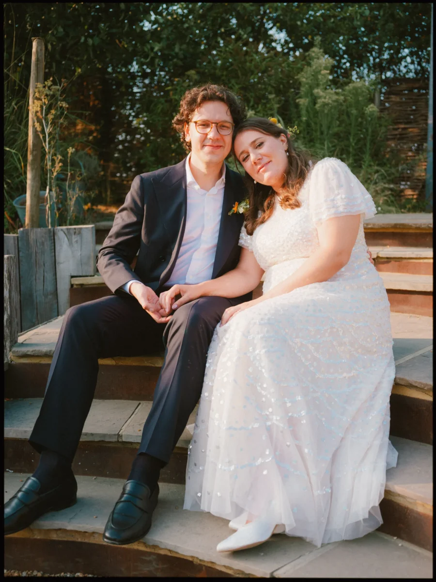 Maya rests her head on Lukas's shoulder as they sit hand in hand on stone garden steps in warm evening light
