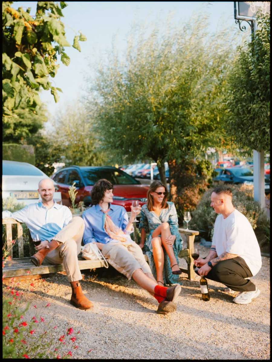 Four guests relax with wine on a wooden bench and gravel in the sunshine, chatting and laughing outside The Bell