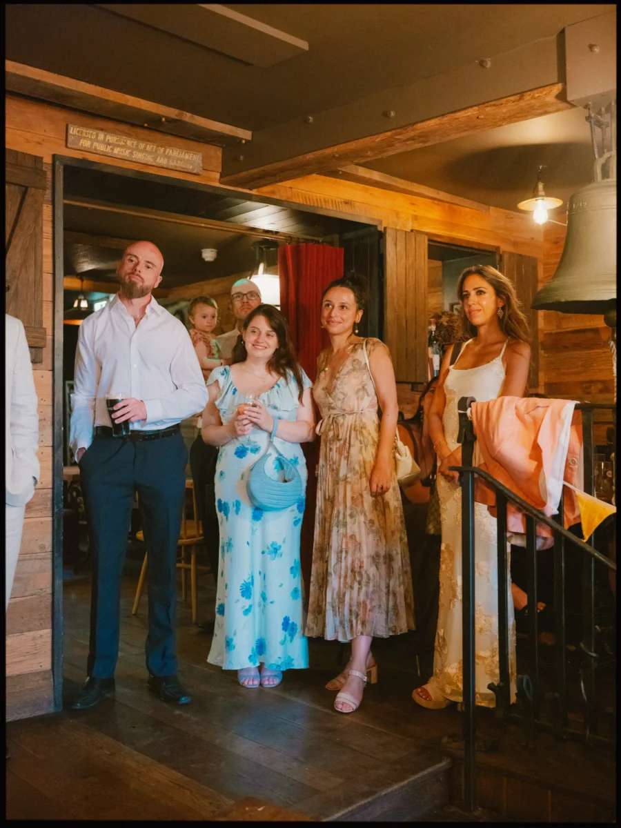 Wedding guests in summer dresses stand smiling and watching, one man holding a pint, beside the big bell inside The Bell
