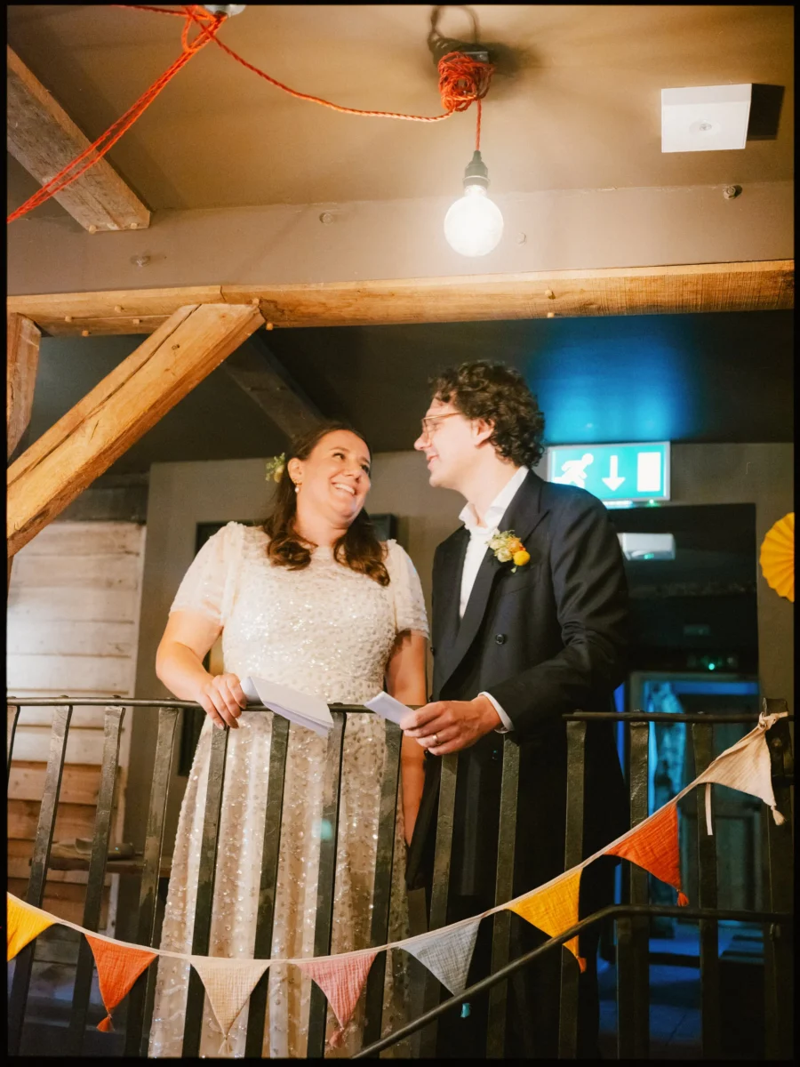 Maya and Lukas laugh together holding folded notes at a balcony railing, bunting strung below, inside The Bell