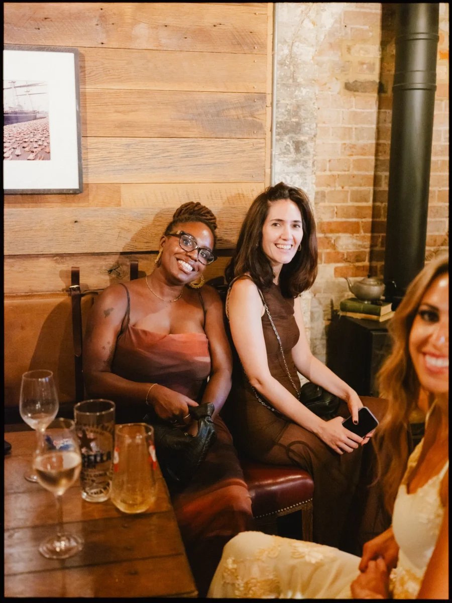 Two guests in brown dresses smile from a leather banquette beside exposed brick at The Bell, drinks on the table