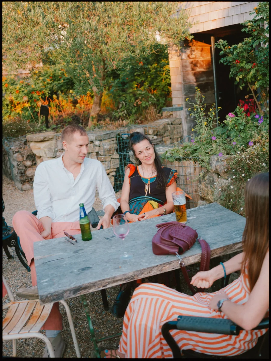 Guests chat around a weathered wooden table with a beer and wine in The Bell's garden, golden evening light