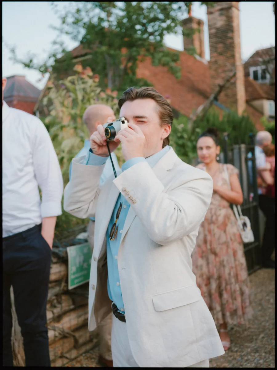 Guest in a cream linen blazer and blue shirt squints as he takes a film camera photo outside The Bell, guests behind