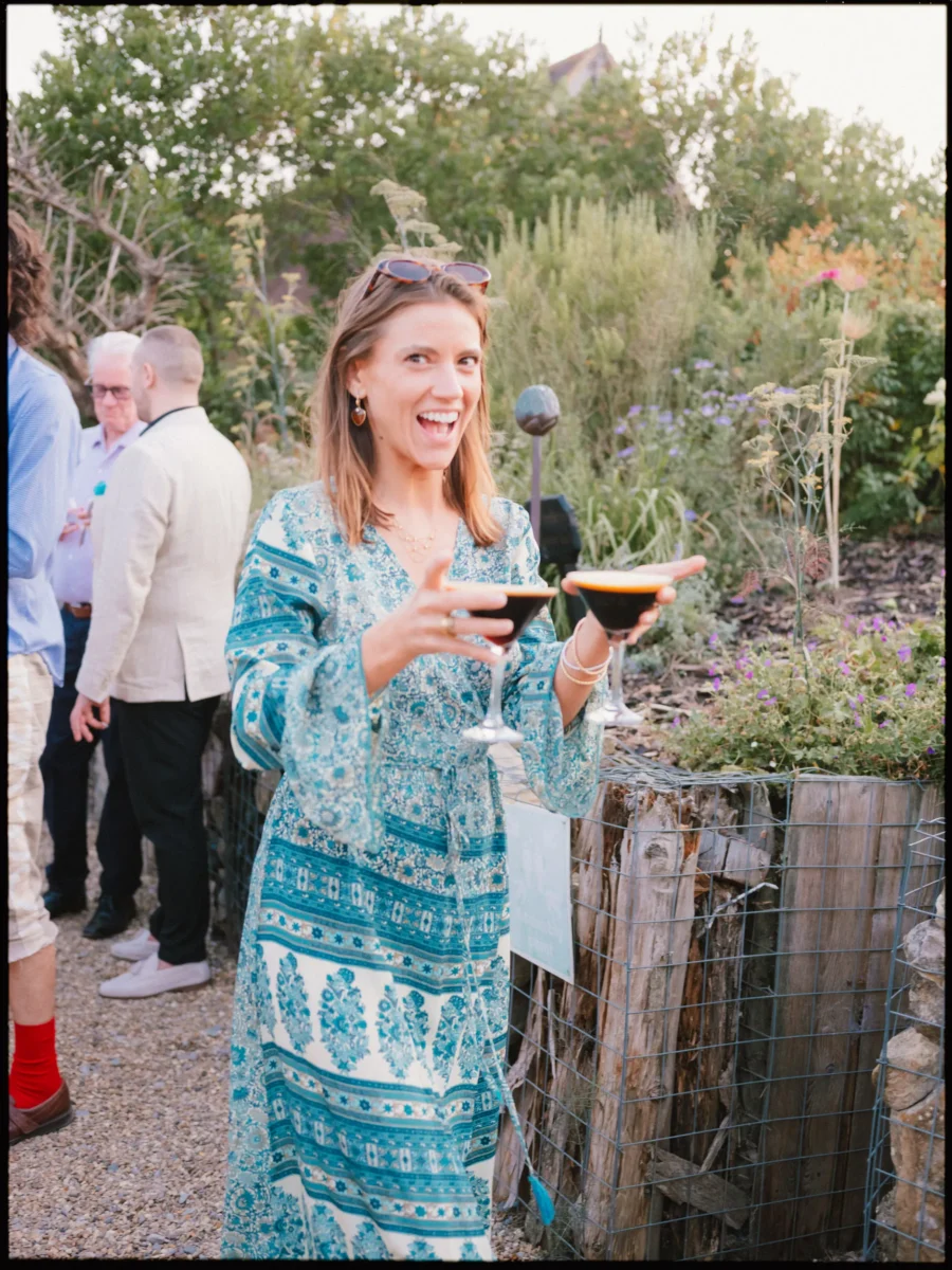 Guest in a teal print dress grins at the camera holding two espresso martinis in The Bell's garden