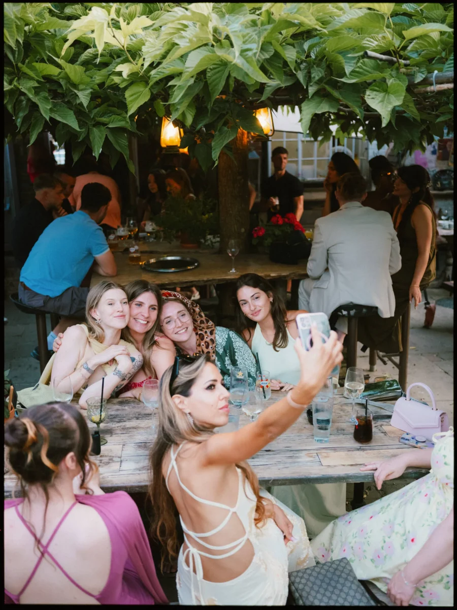 Woman in a white strappy dress takes a selfie with smiling friends at a rustic outdoor table under leafy vines