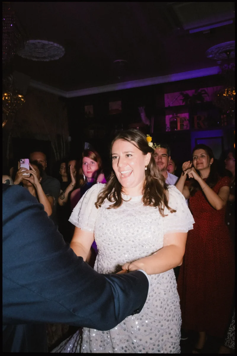 Maya laughs mid-dance in a sparkly white dress as guests clap and film on their phones in the purple-lit room