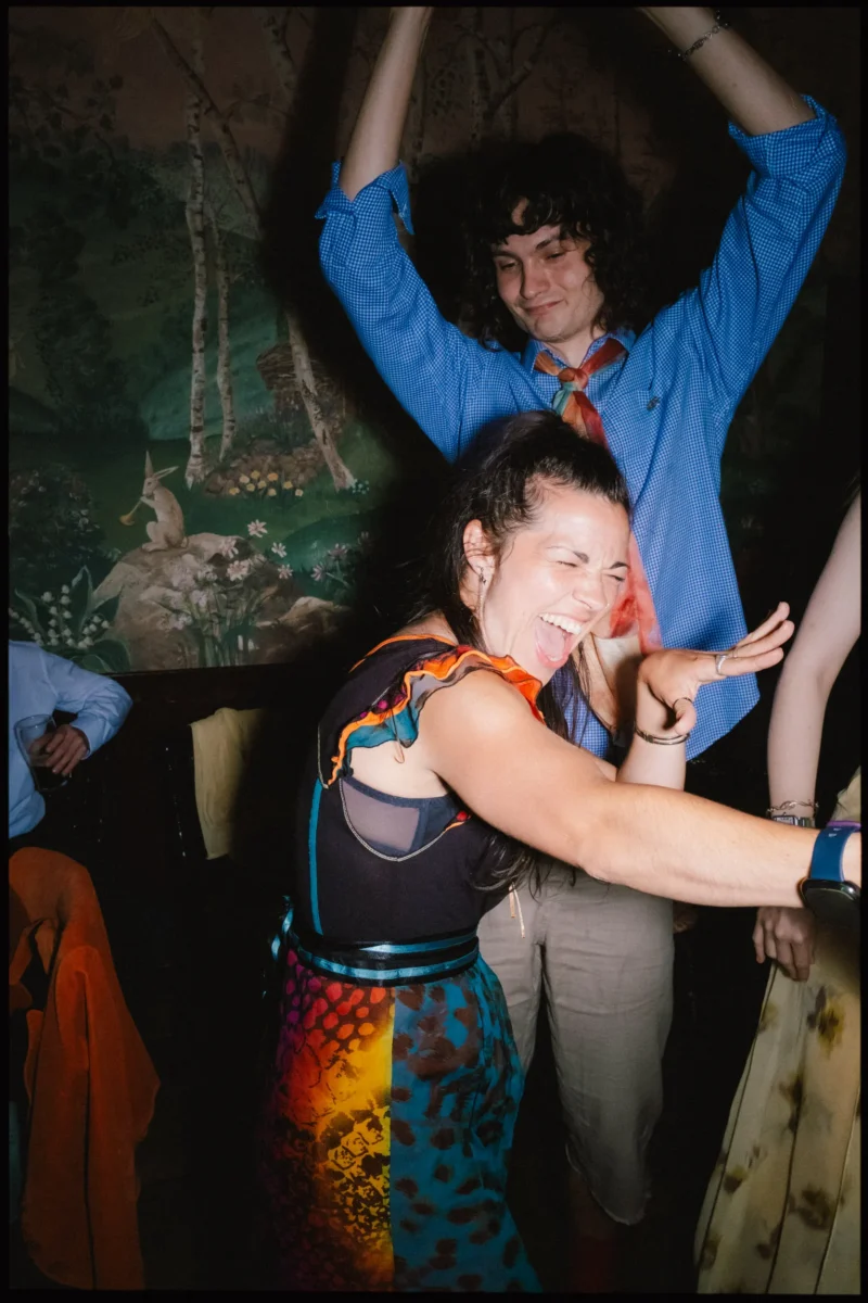 Woman in a colourful patterned skirt laughs with eyes shut while dancing, a man with arms raised behind her, at The Bell