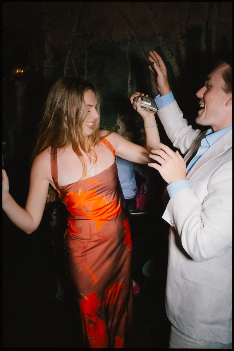 Guest in a burnt-orange slip dress dances with a laughing man in a cream blazer, who holds a small camera, at The Bell
