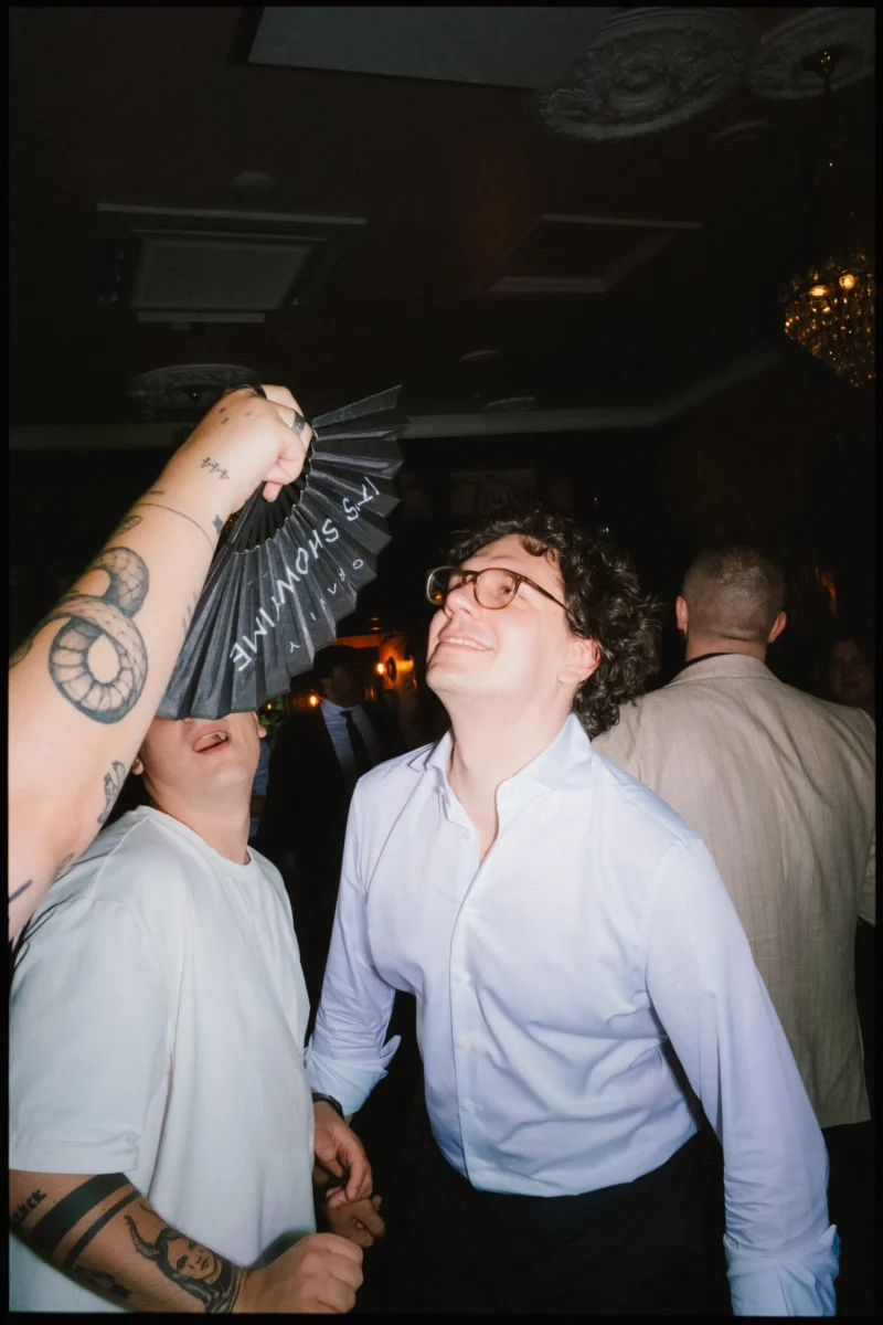 Guest in glasses and white shirt grins upwards as a tattooed arm holds a black fan over a friend's face on the dance floor