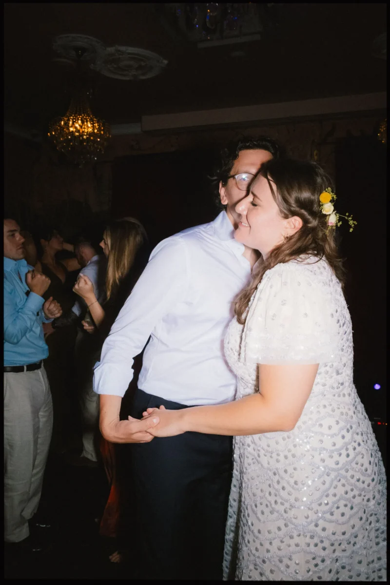 Maya and Lukas sway cheek to cheek, hands clasped, her eyes closed and smiling, as guests dance beneath a chandelier