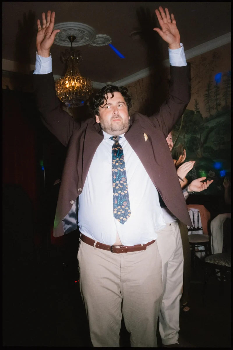 Guest in a brown blazer and floral tie throws both hands in the air on the dance floor beneath a chandelier at The Bell