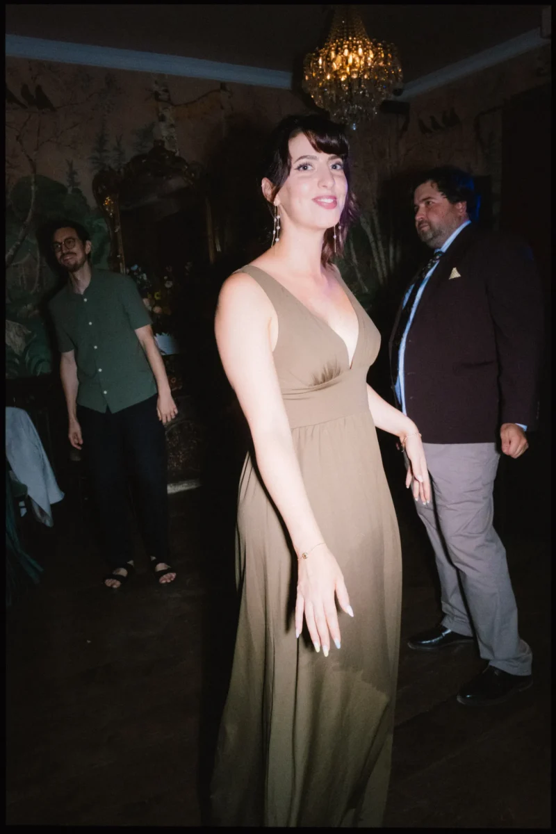 Guest in an olive satin gown smiles at the camera mid-dance as two men look on, under a chandelier at The Bell