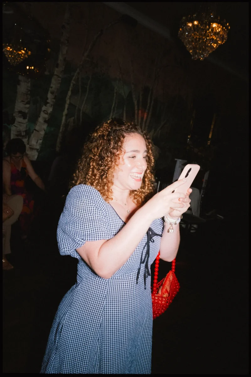 Guest in a blue gingham dress laughs while looking at her phone, red beaded bag at her side, birch wallpaper at The Bell