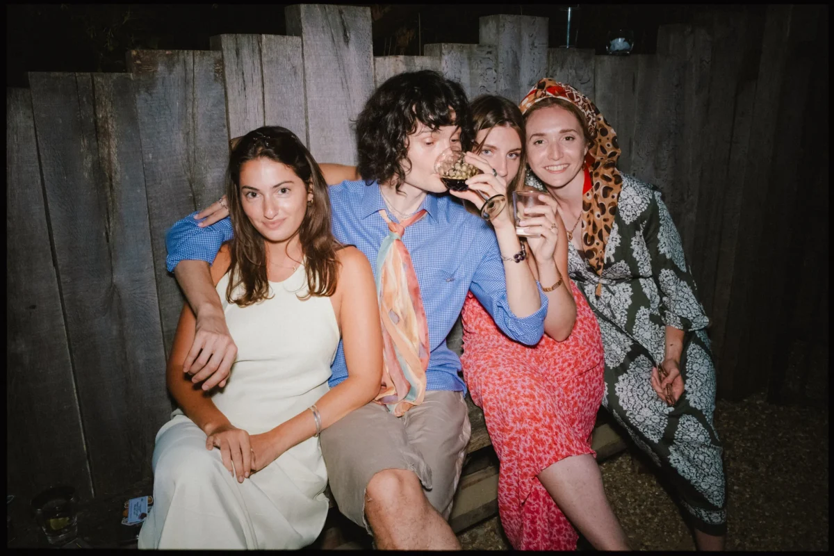 Four guests squeezed onto a bench at night, one sipping a drink, in front of a weathered wooden fence at the wedding