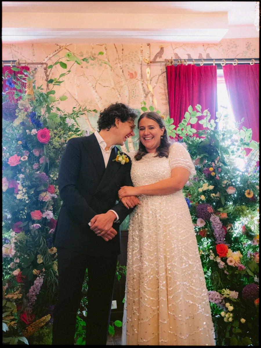 Maya and Lukas laugh arm in arm in front of wild flowers, birch-tree wallpaper and magenta curtains at The Bell