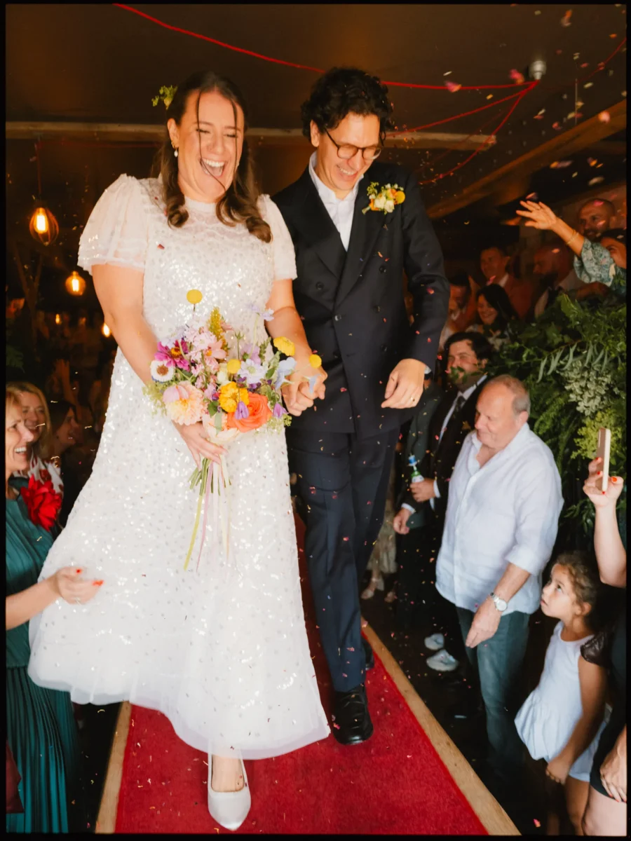 Bride and groom laugh as confetti falls on the red carpet at The Bell in Ticehurst, guests cheering either side