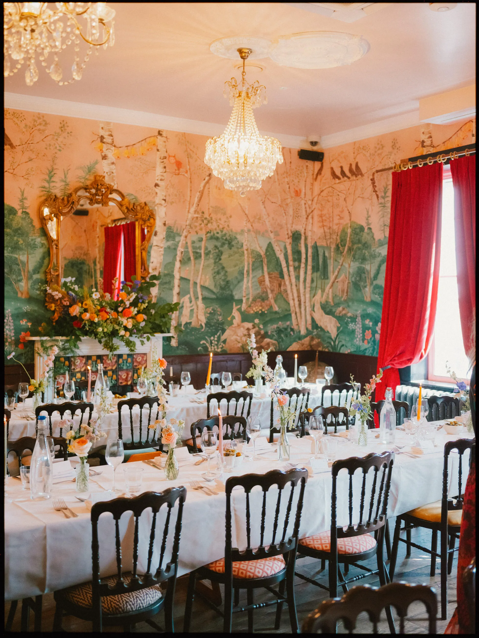 Long wedding tables set with wildflower posies and candles beneath chandeliers, woodland wallpaper behind at The Bell