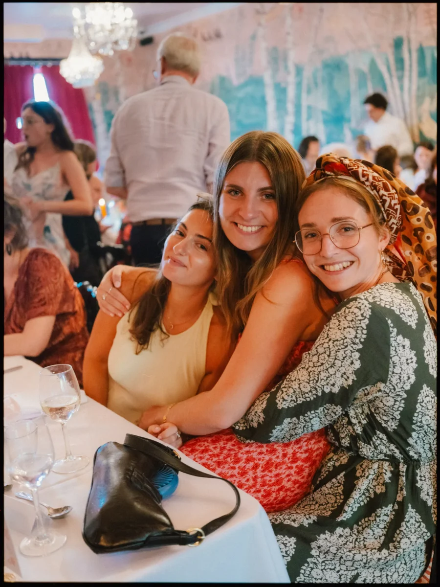 Three women squeezed together and smiling at a wedding table inside The Bell in Ticehurst, guests chatting behind