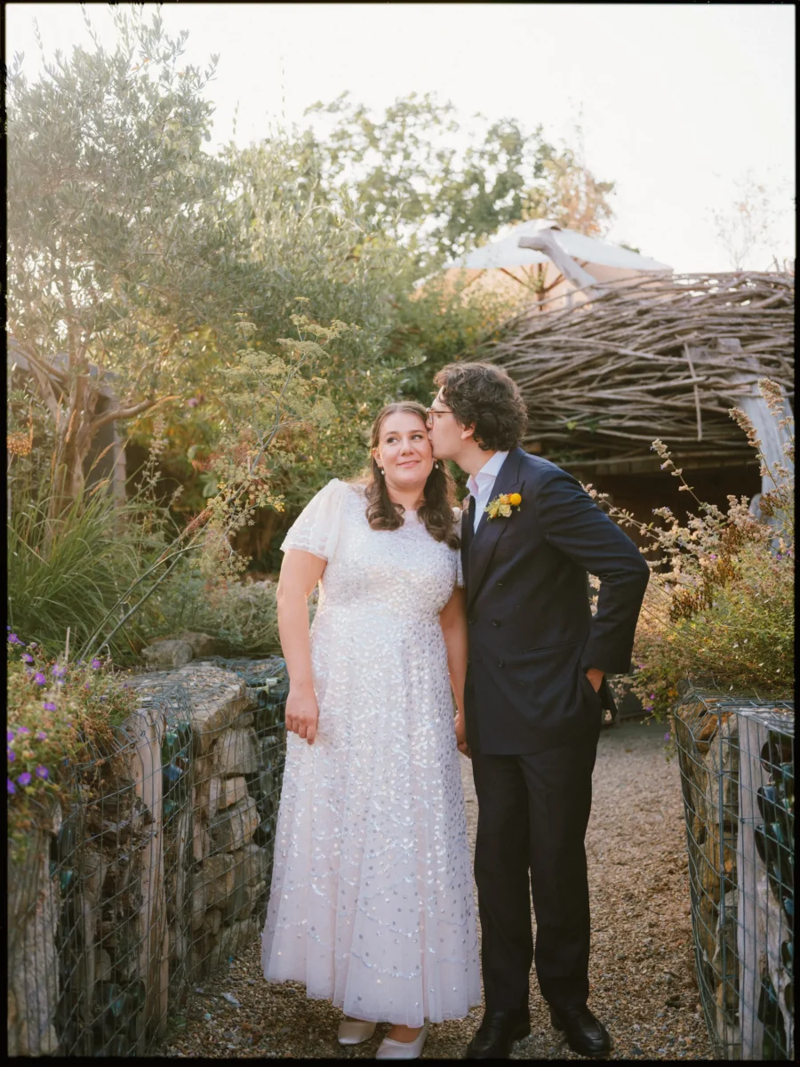 Lukas kisses Maya's temple as she smiles in her sequinned dress, in the evening light of The Bell's garden