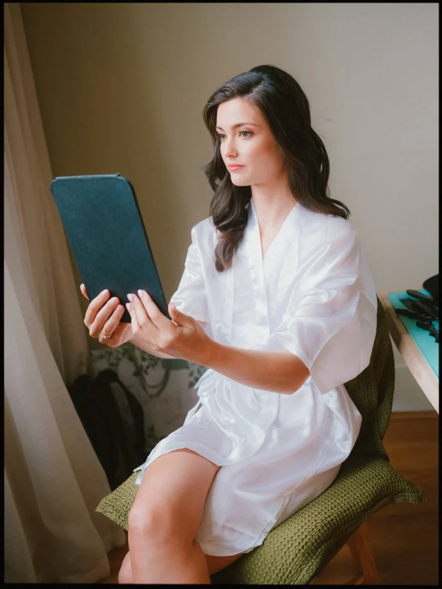 Bride in a white satin robe sits in a green chair, quietly looking at a tablet in her room at the Town Hall Hotel