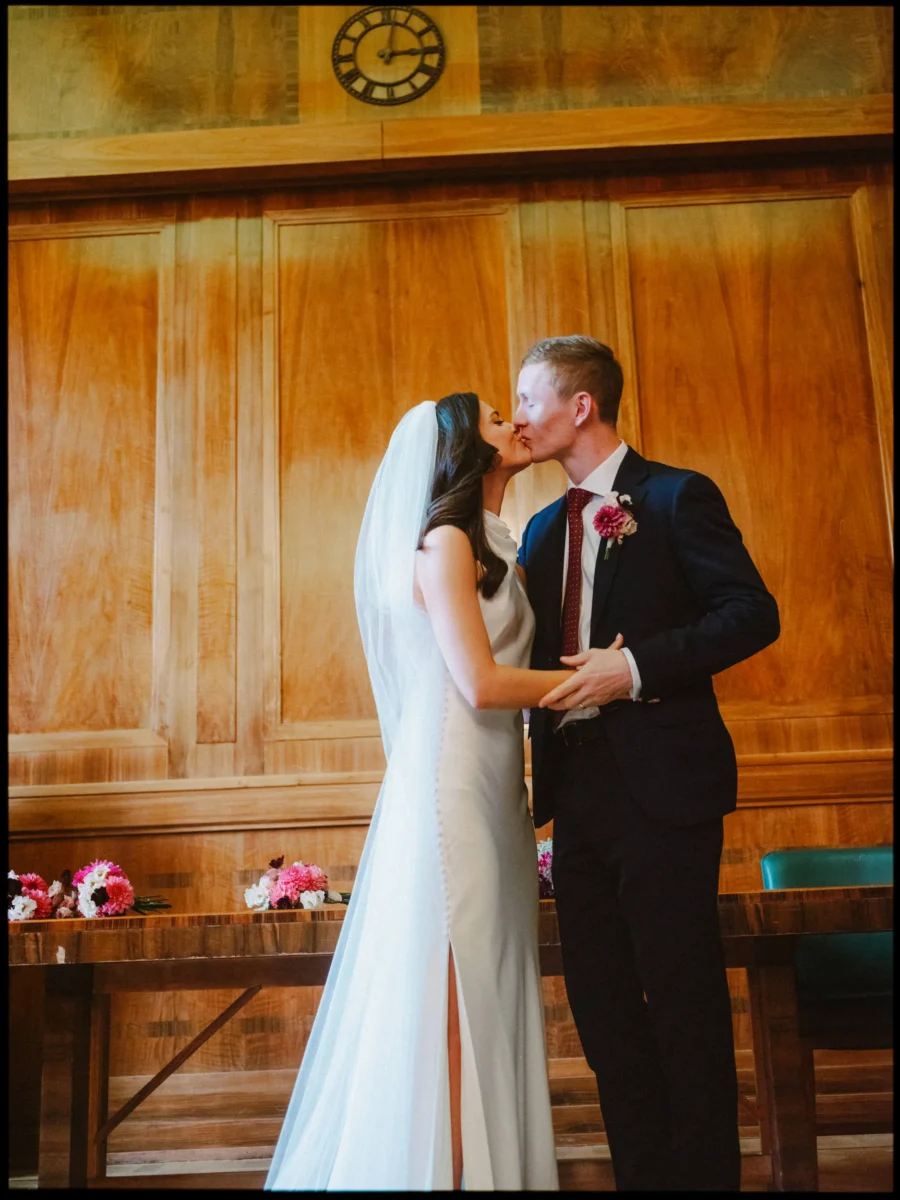 Bride in a veil and groom in a navy suit share a kiss in front of the warm wood panelling of Hackney Town Hall