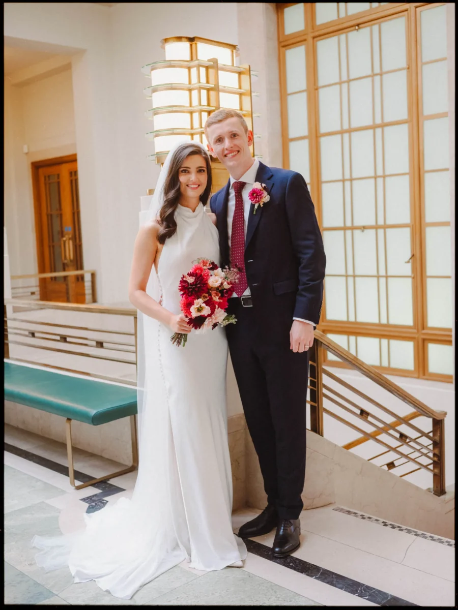 Bride with a red and pink bouquet and groom in a navy suit smile together on the art deco stairs at Hackney Town Hall