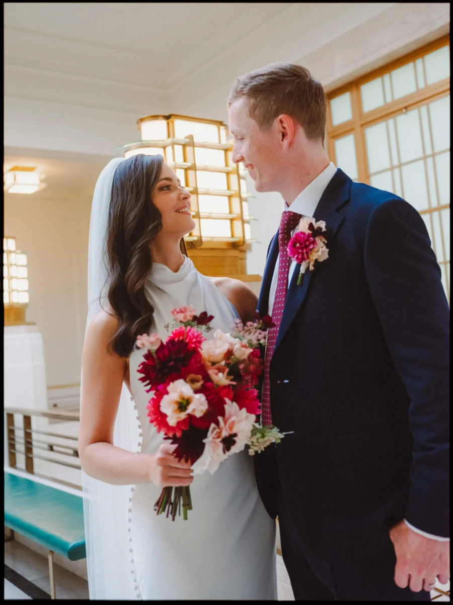 Bride in a veil and cowl-neck gown smiles up at her groom beside glowing amber lamps in Hackney Town Hall