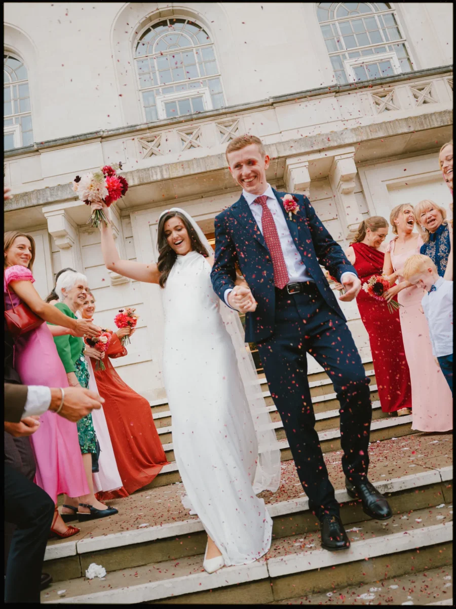 Grinning groom leads his bride by the hand down the Hackney Town Hall steps past guests in bright dresses