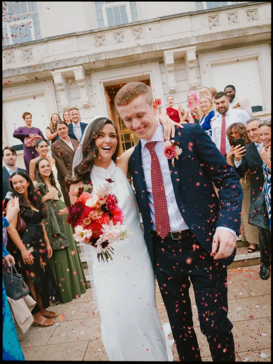Bride and groom laugh as guests throw confetti on the steps of Hackney Town Hall, bright dahlia bouquet in hand