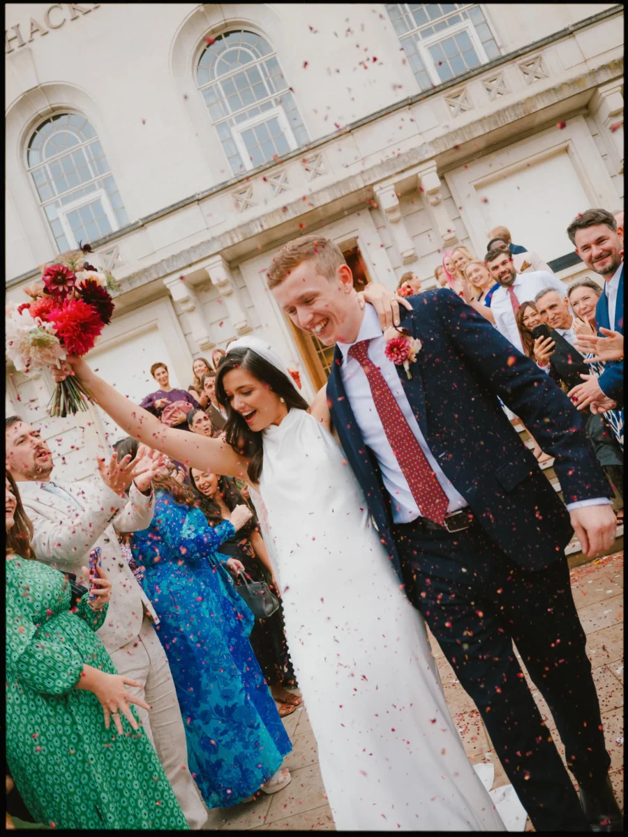 Couple dash through a shower of confetti, bride raising her red dahlia bouquet high as cheering guests line the steps