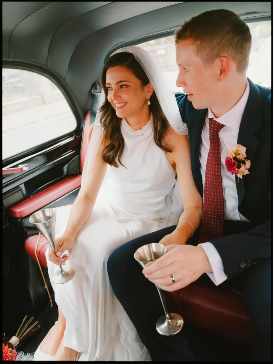 Bride and groom sit close in the back of a black taxi, each holding a silver champagne flute and smiling out the window