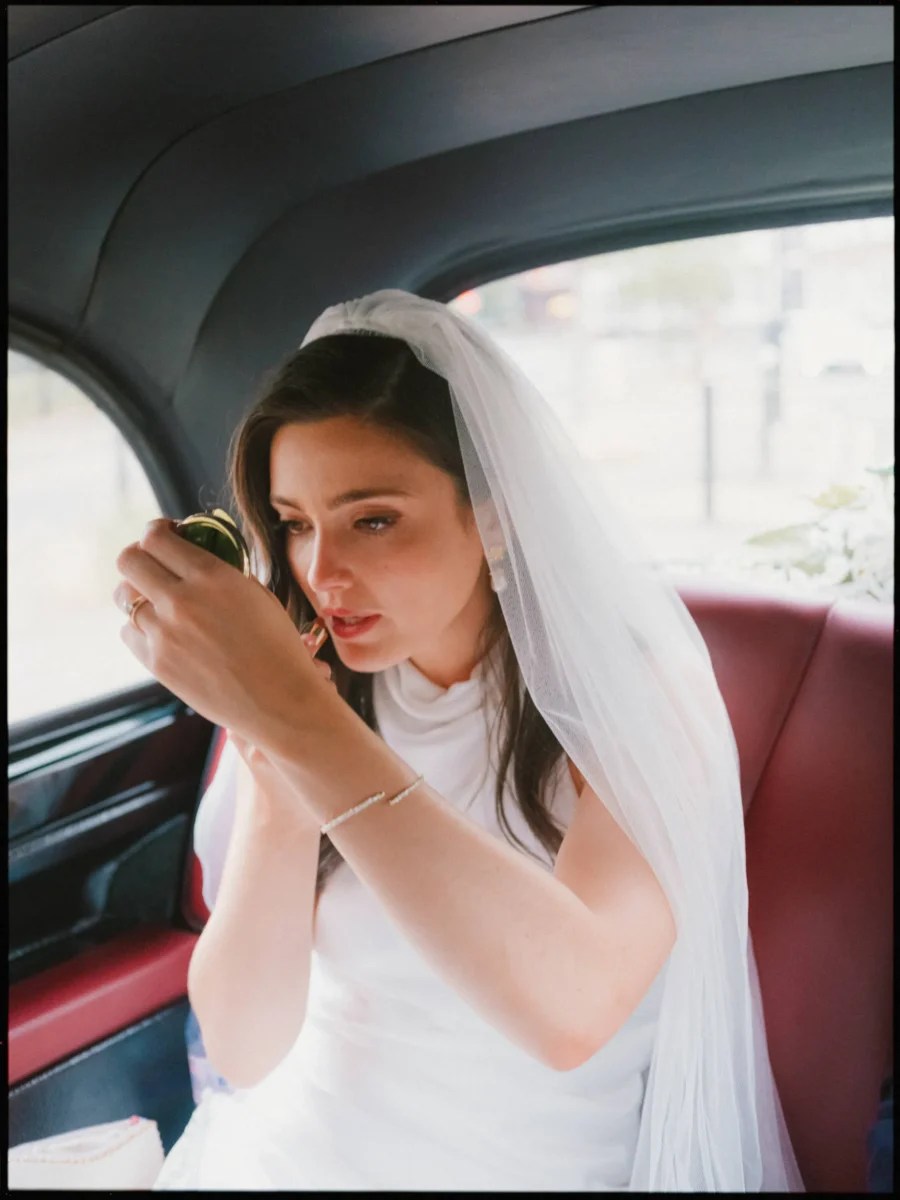 Bride in a veil checks her make-up in a small compact mirror in the back of a taxi with red leather seats