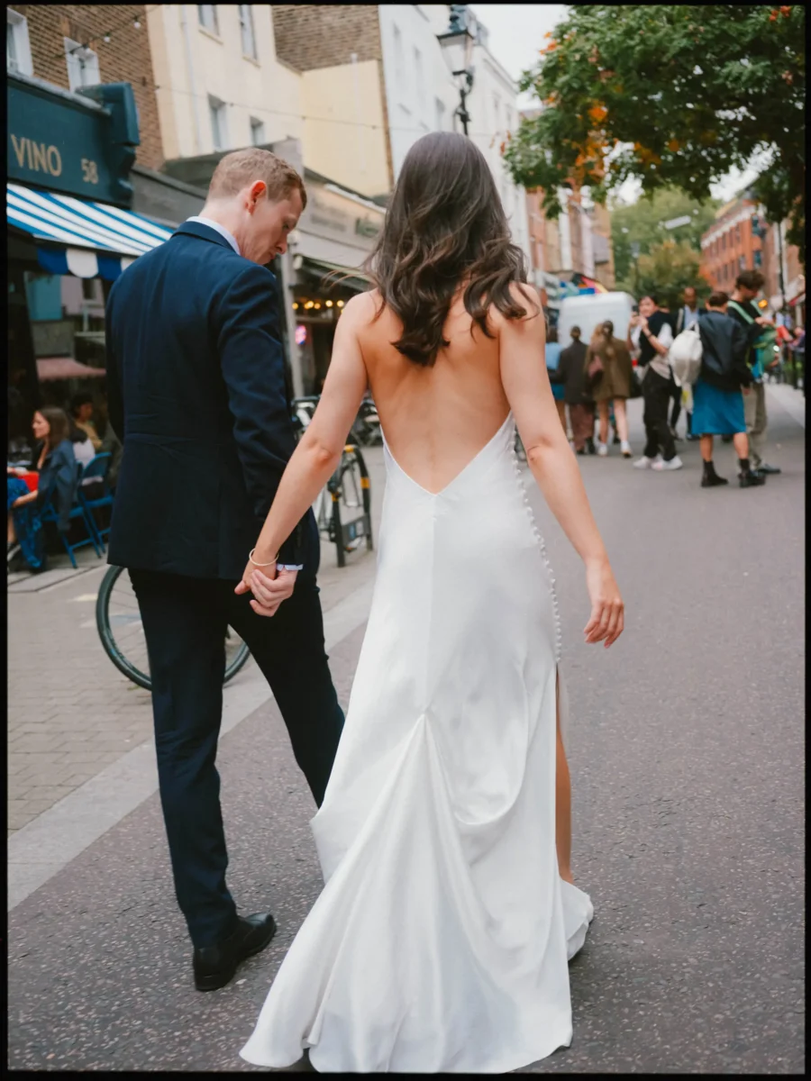 Bride in a low-back white satin gown and groom in navy walk hand in hand down a busy London street past cafe tables