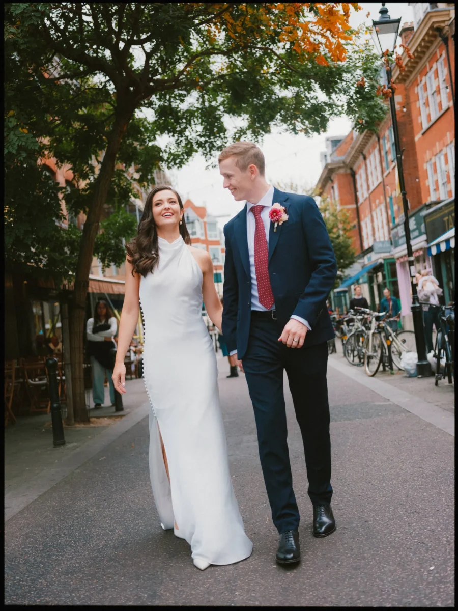 Bride in a white halterneck gown and groom in a navy suit walk laughing down a tree-lined street, past bikes and cafés