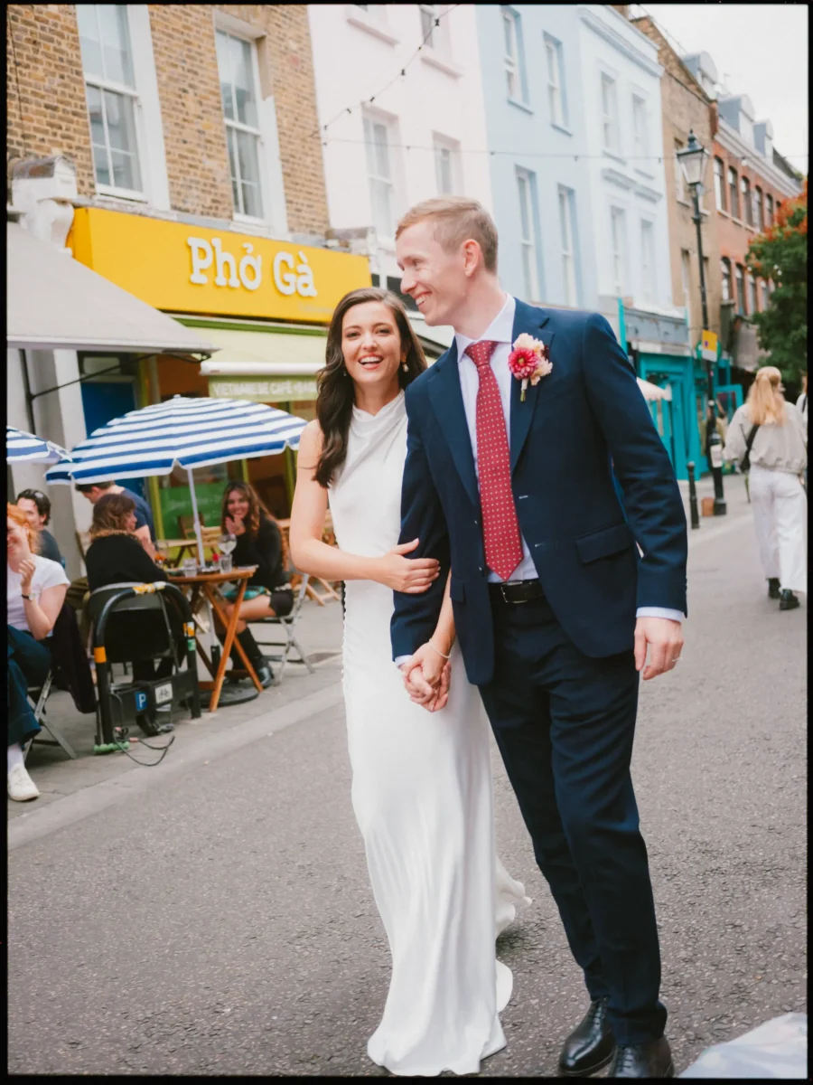 Laughing bride and groom in a red tie stroll arm in arm past diners outside a yellow Phở Gà café in Exmouth Market