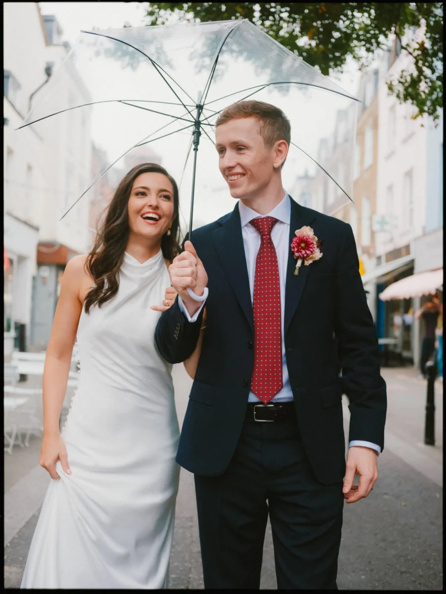 Laughing bride and smiling groom in a red dahlia buttonhole share a clear umbrella on a street in Exmouth Market