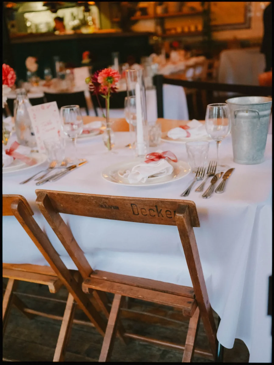 Wedding table set with red-ribboned napkins, a dahlia in a bud vase and worn wooden folding chairs at Coin Laundry