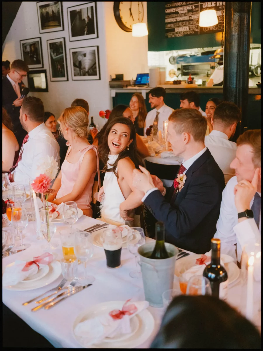 Bride in a white halterneck top turns laughing to her groom at a candlelit dinner table full of guests at Coin Laundry
