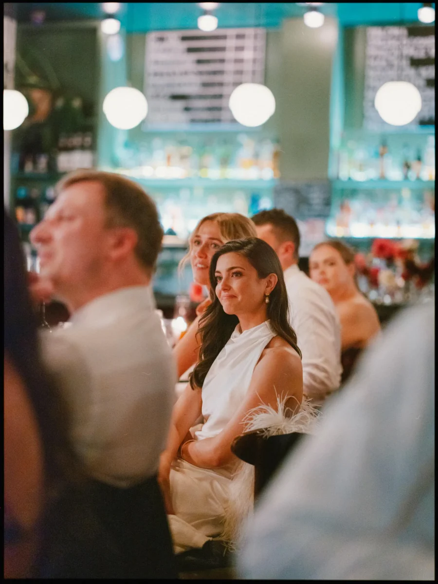 Bride in a white halterneck top and feathered skirt listens with a soft smile among dinner guests at Coin Laundry