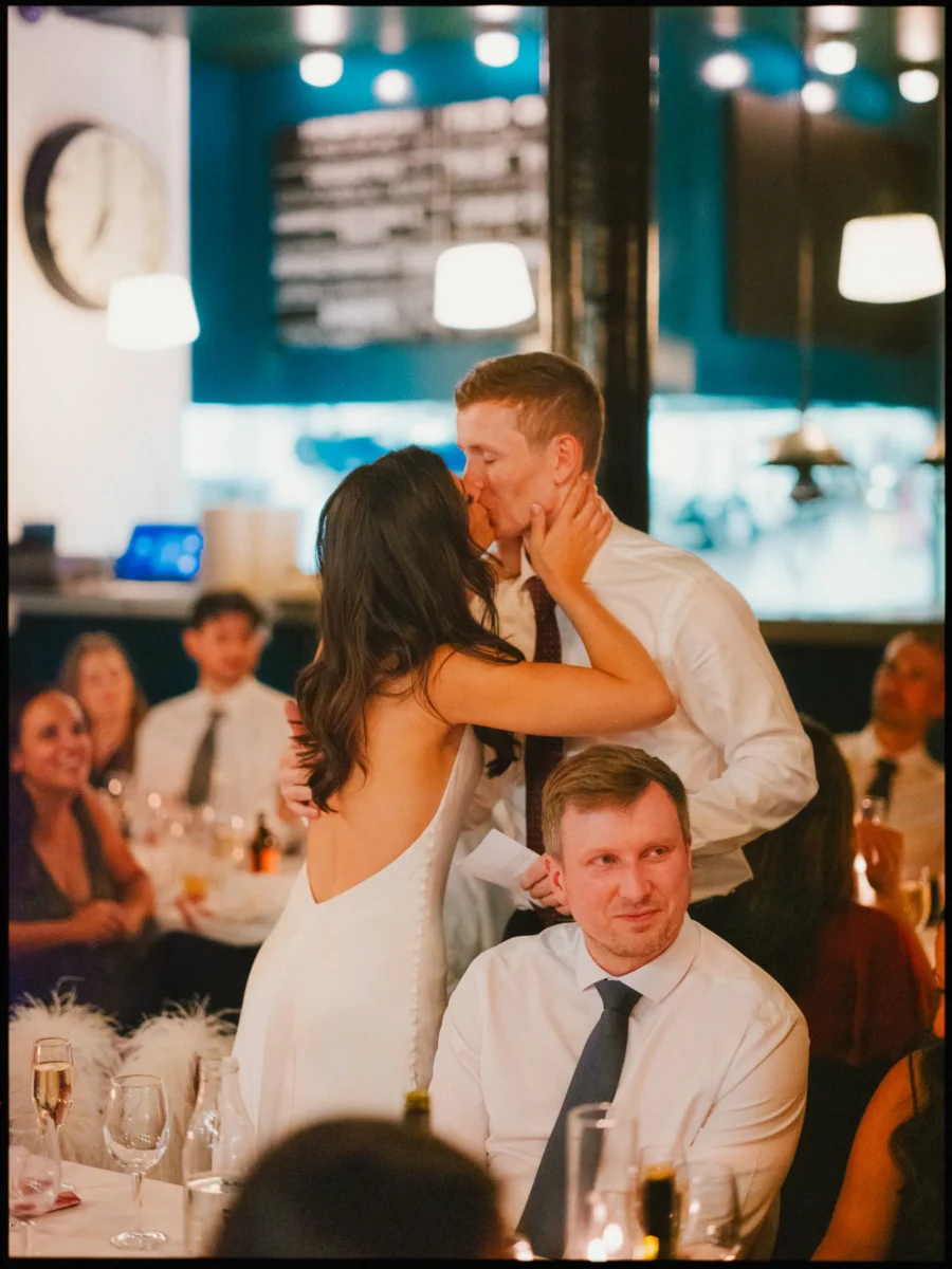 Bride in a backless white gown kisses her standing groom at dinner in Coin Laundry as a seated guest grins on
