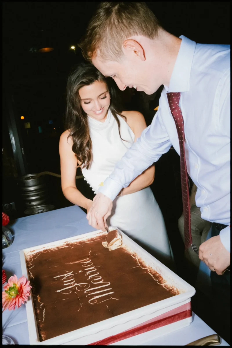 Smiling bride watches as her groom in a red tie lifts the first slice of tiramisu, cutting the cake at Coin Laundry