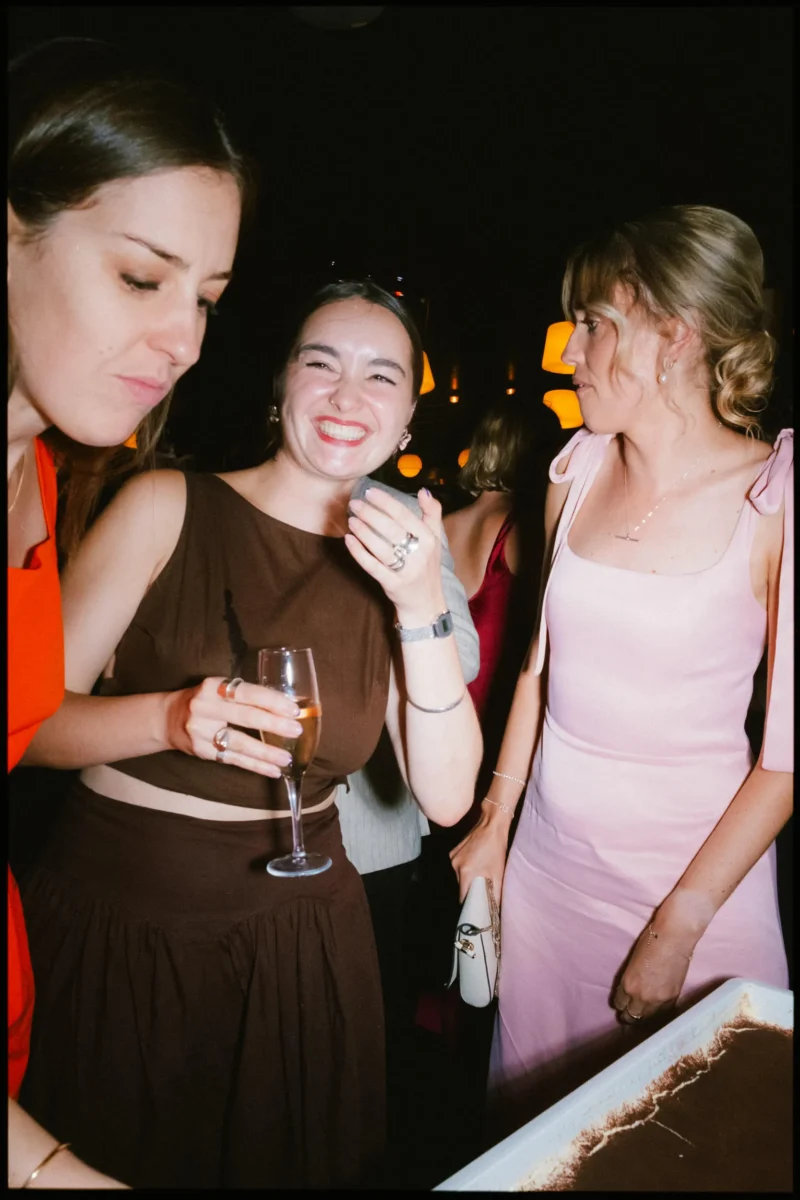 Guest in a brown top laughs with a champagne flute as friends lean in towards the tiramisu in the dark Coin Laundry basement