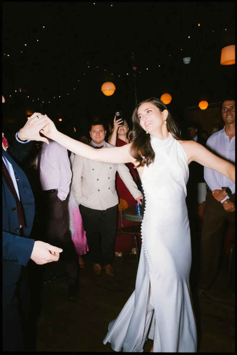 Bride in a white satin halterneck gown spins with arms outstretched as guests watch from the Coin Laundry dance floor