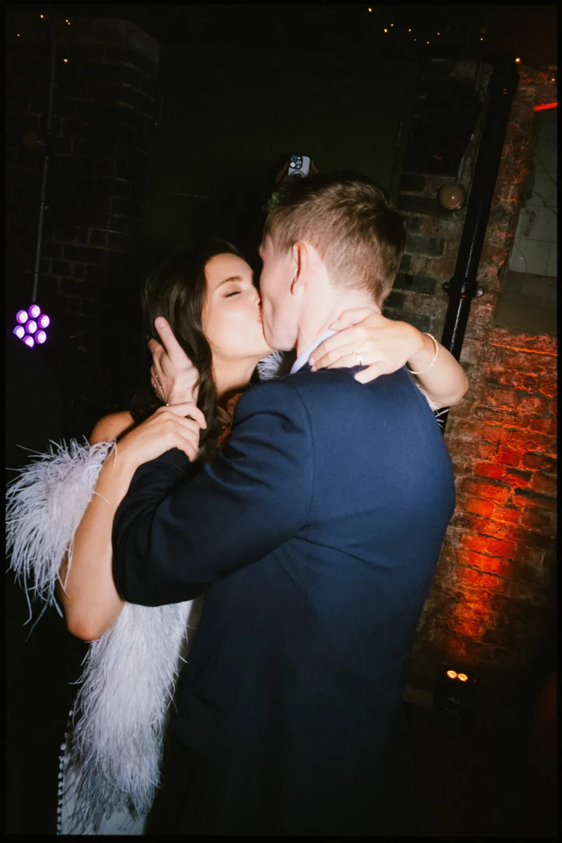 Bride in a white feathered stole wraps her arms around her groom in a deep kiss against exposed brick at Coin Laundry