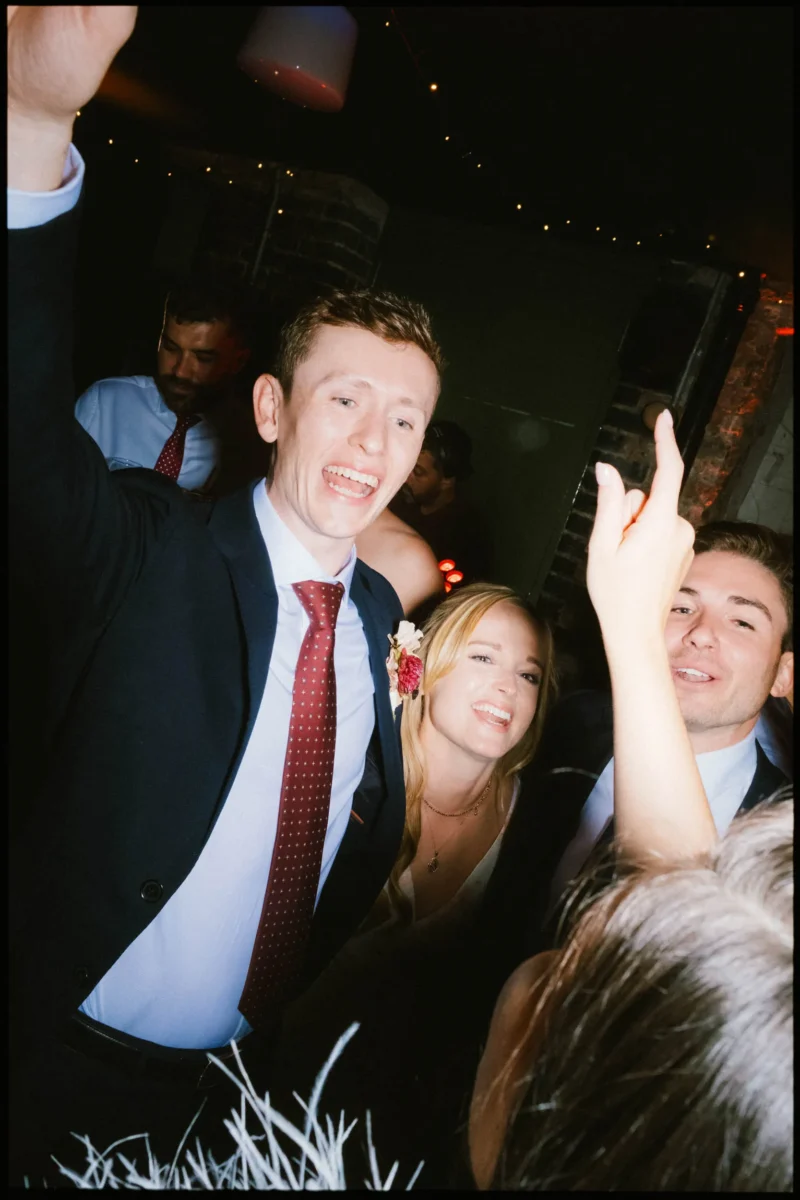 Guest in a red tie sings with arm raised as a smiling blonde woman and friends crowd in on the Coin Laundry dance floor
