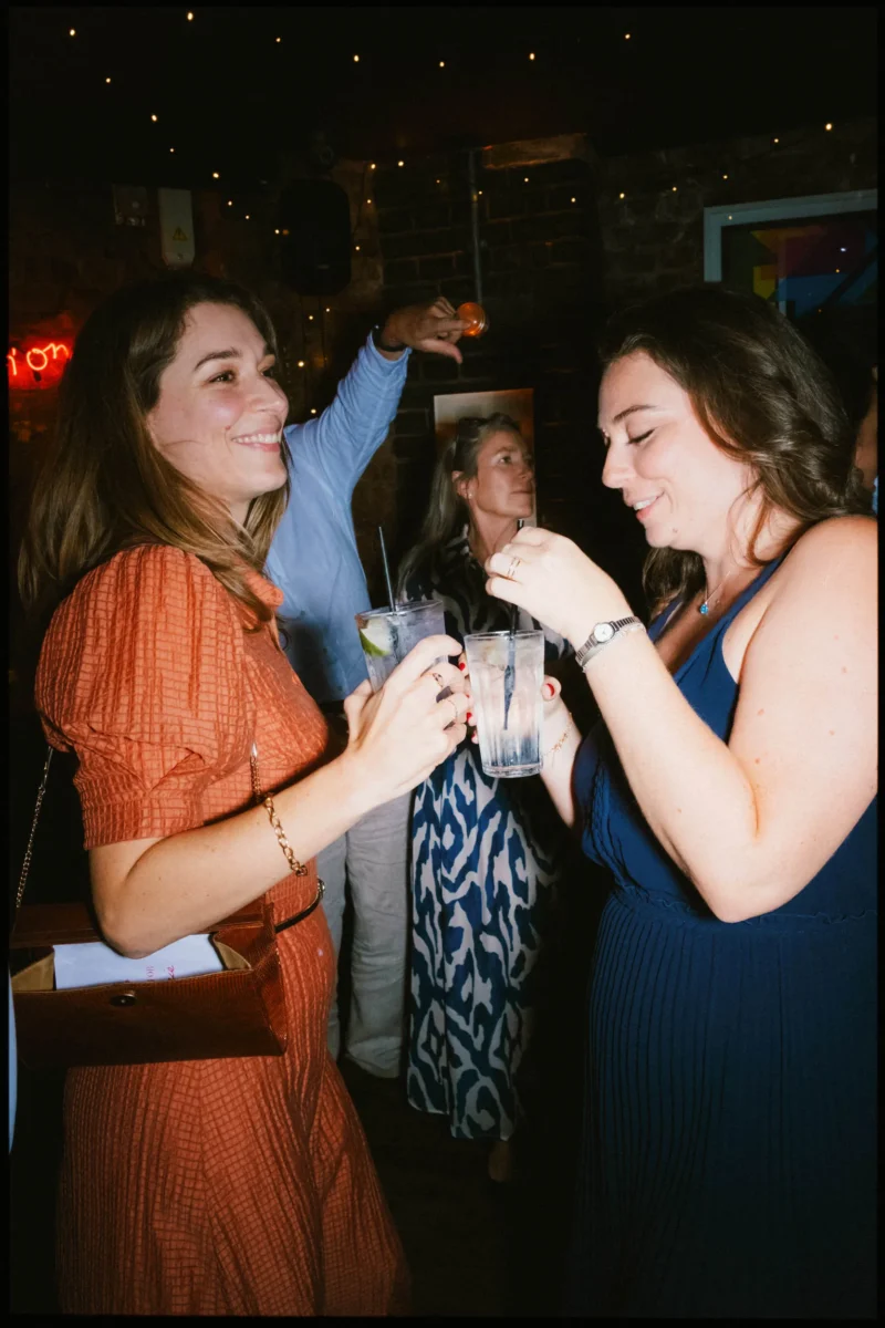 Two women clink glasses and laugh together in the dimly lit Coin Laundry basement, fairy lights glowing behind them
