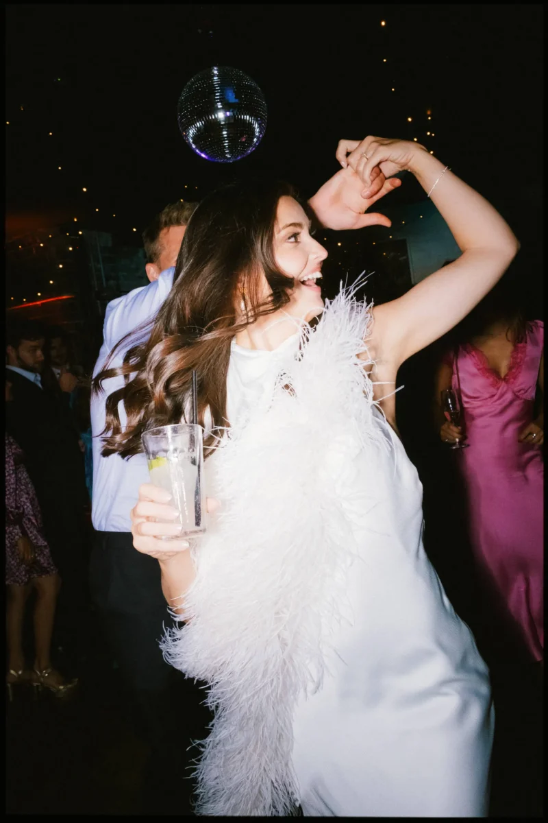 Bride with long hair swinging dances, arm raised and drink in hand, under a disco ball at Coin Laundry