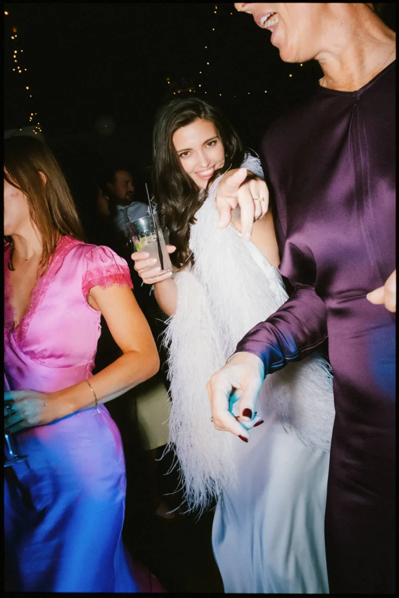 Bride in a white feathered stole points at the camera with a drink in hand, dancing with guests in the Coin Laundry basement