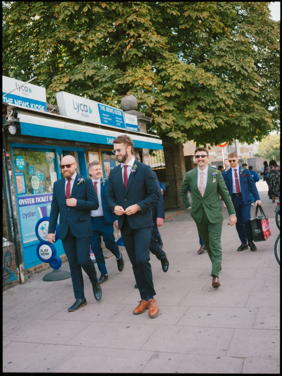 Groom and groomsmen in navy and green suits walking along a Bow pavement past a news kiosk, laughing and relaxed